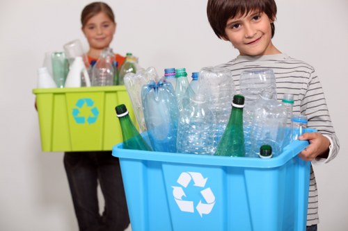 A young boy with dark brown hair, wearing a grey and white striped long-sleeve shirt, is holding a large blue recycling bin filled with various empty plastic bottles, including clear, green, and light blue containers, some with caps and labels still attached. Behind him, a young girl with light brown hair, dressed in a grey top, stands holding a smaller green recycling bin containing additional plastic bottles and containers. Both children are positioned indoors against a plain, light-colored background, possibly in a home or storage area, illustrating waste sorting or recycling activities. The blue bin prominently features a white recycling symbol, indicating it is designated for plastic waste. The scene emphasizes environmentally conscious waste separation, with the children engaged in recycling practice consistent with rubbish management services in the Shortlands area, underlining the importance of responsible disposal. The environmental context and the focus on plastic waste collection highlight the relevance to local rubbish removal and recycling efforts facilitated by Garden Clearance Shortlands.