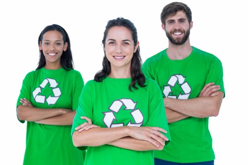 A group of three individuals standing outdoors in front of a plain background, each wearing bright green T-shirts featuring a white recycling symbol. The person in the center, a woman with dark hair styled in loose waves, is smiling confidently with arms crossed. To her left, another woman with long dark hair is also smiling and has her arms crossed, while to her right, a man with a beard and short dark hair is smiling broadly, with his arms crossed as well. The setting suggests a team involved in environmental or waste management activities, possibly representing a local rubbish removal service such as Garden Clearance Shortlands. The focus is on their cheerful, professional appearance and the universal recycling symbol on their shirts, highlighting their commitment to eco-friendly waste disposal and garden clearance services in the Shortlands area, within the Greater London region. The overall atmosphere is positive, professional, and environmentally conscious, fitting for a company specializing in rubbish removal services in the local vicinity.