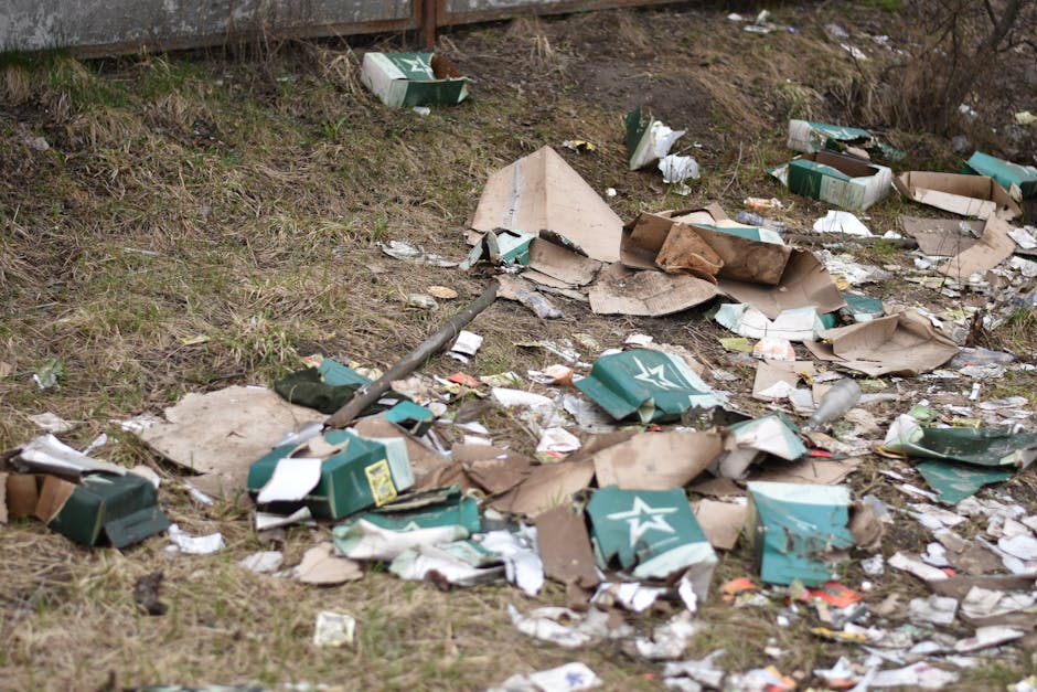 A photograph depicting a collection of various discarded waste items placed on an outdoor paved driveway in a residential area. In the foreground, there are several large black plastic trash bags fill