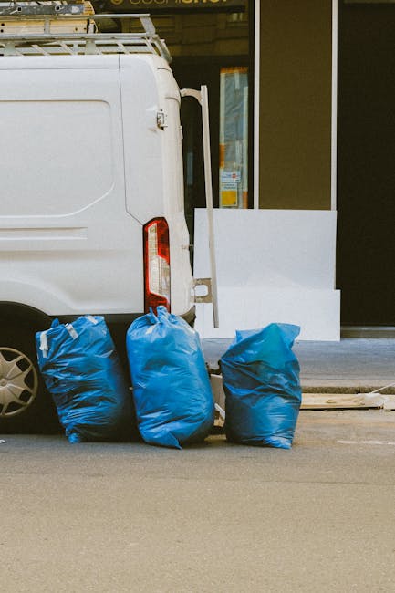 The image shows the rear section of a white commercial van parked alongside a building with large glass windows and a recessed entrance. Three large blue plastic rubbish bags are positioned on the pav