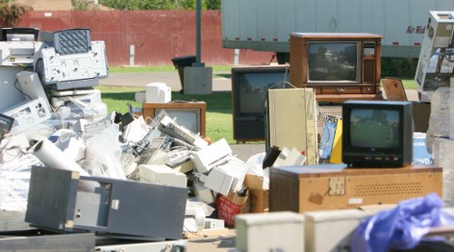A large pile of discarded electronic items and household appliances, including several old CRT televisions with wooden and plastic casings, computer monitors, keyboards, and miscellaneous electronic waste, all stacked and scattered on a paved outdoor area. The background features a red brick wall, green grass, and a bus stop shelter with a bench, indicating a typical urban environment. The electronic waste appears weathered, with some units showing signs of age such as scratches and faded screens. This scene exemplifies clutter associated with rubbish removal services provided by Garden Clearance Shortlands in the local area, likely within or near the postcode region of Shortlands, emphasizing a professional collection and disposal of bulky electronic waste.